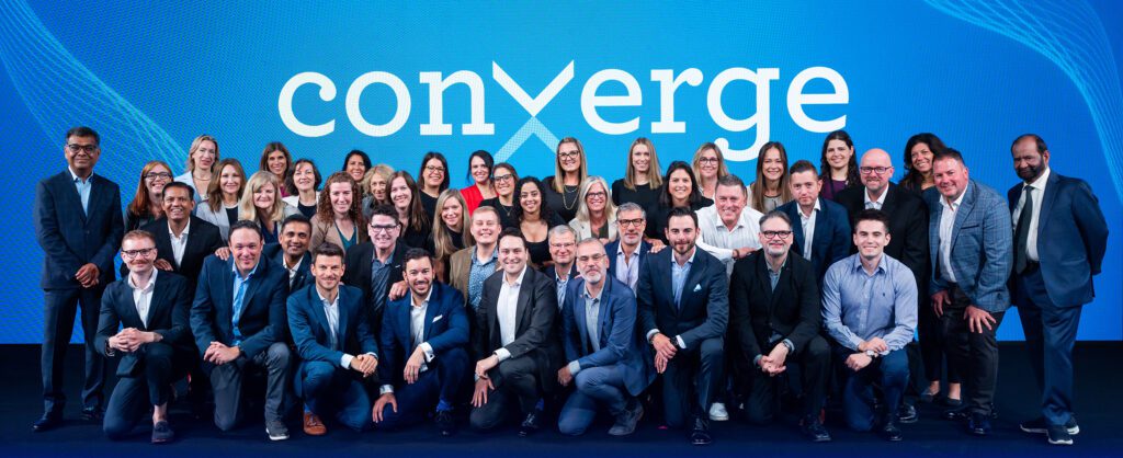 Group photo of IDeaS employees gathered on stage in front of a large Converge 2025 banner