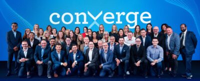 Group photo of IDeaS employees gathered on stage in front of a large Converge 2025 banner
